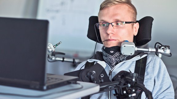 Foto eines Mannes mit blonden Haaren, der im Rollstuhl sitzt und mittels Mundstab und Trackball Maus versucht einen Computer zu bedienen.